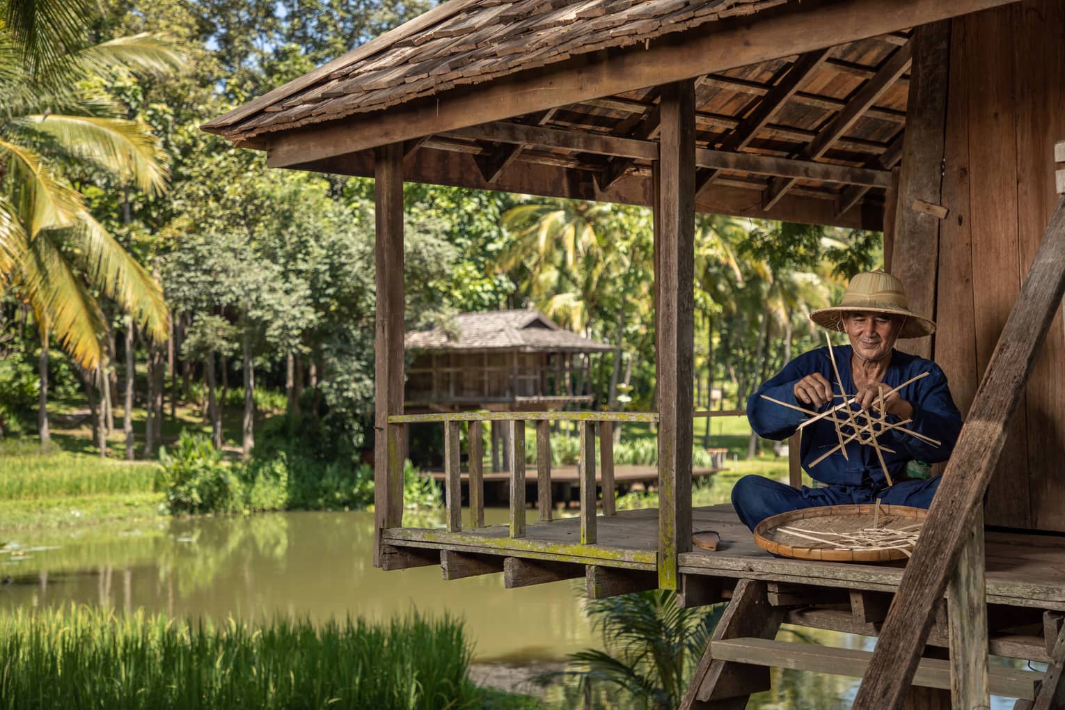 Man weaves a traditional Thai basket on the porch of a riverside pagoda