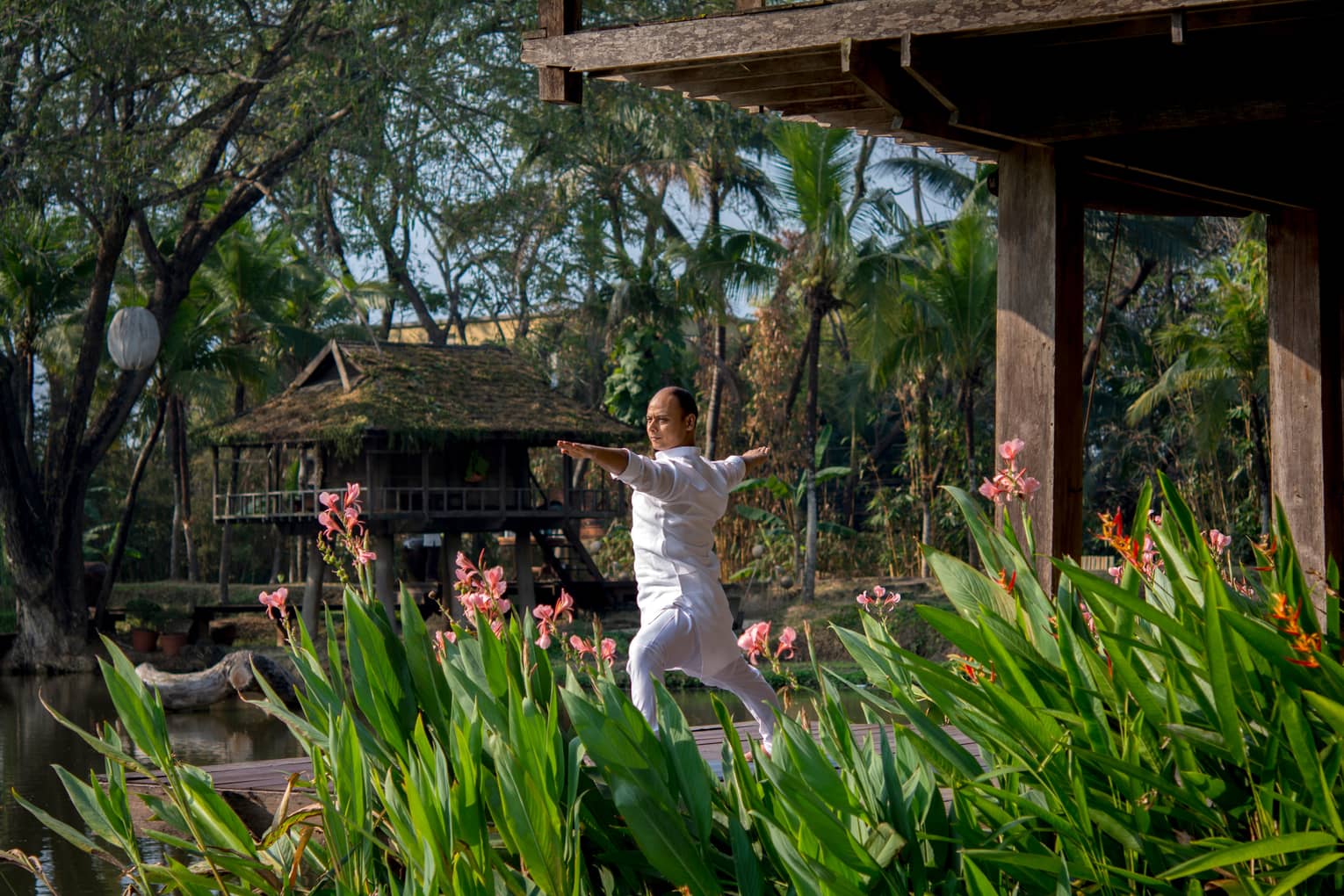 Resident yogi Dheera wearing white in yoga pose with arms outstretched by tall flowers, leaves