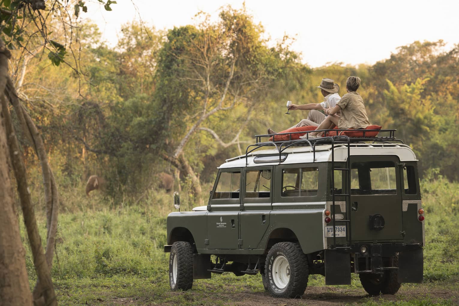 A couple sits on the roof of a Land Rover in the Bush, drinking a cocktail