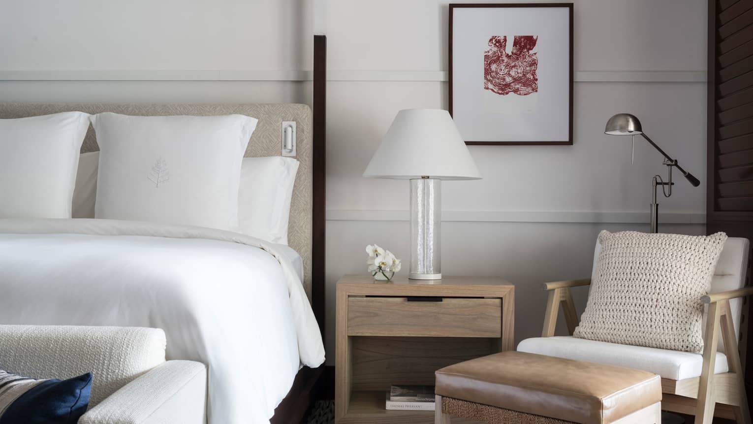 Close-up of white hotel bed, nightstand with white lamp, modern armchair and leather footstool