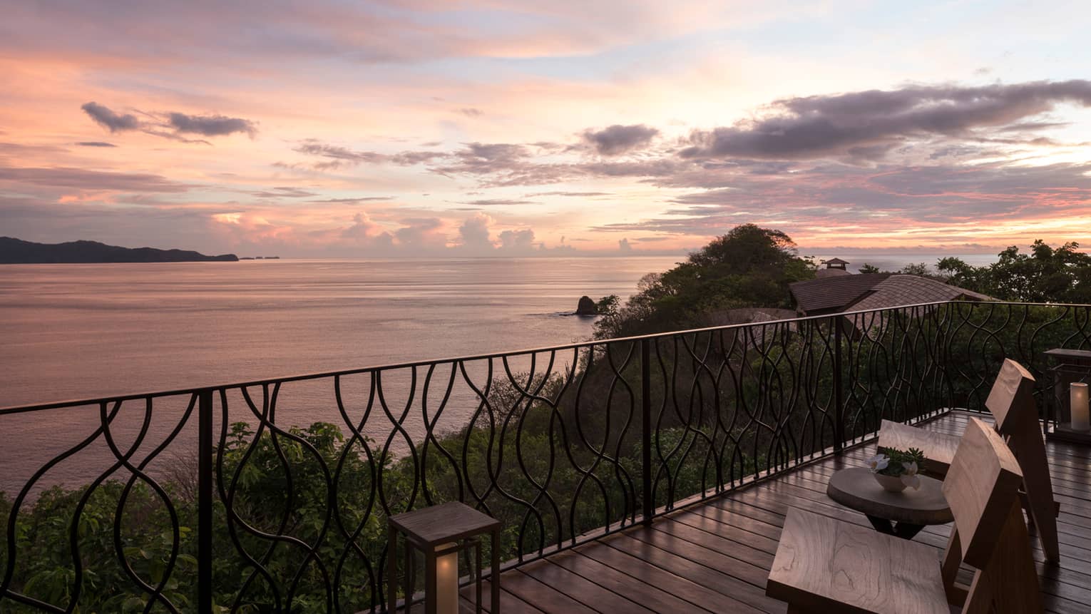 Patio chairs on wood deck with iron balcony at sunset, high above trees and ocean