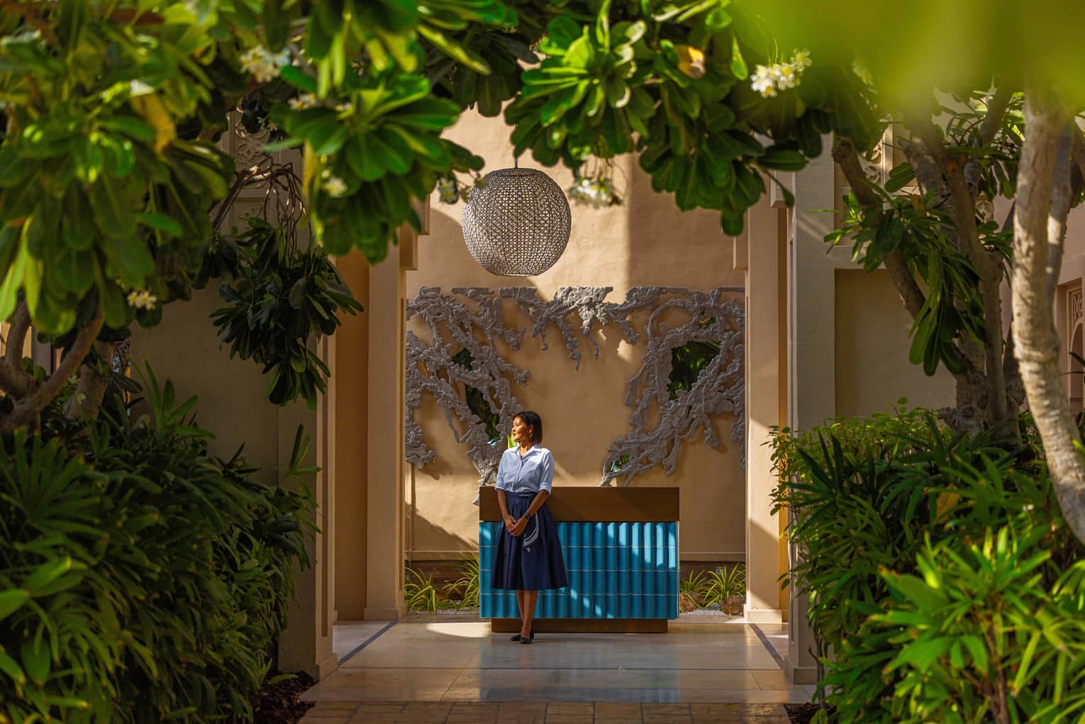 Woman staff member wearing light-blue button down shirt and dark blue knee-length skirt stande next to a blue-tiled counter set surrounded by bright green plants and trees