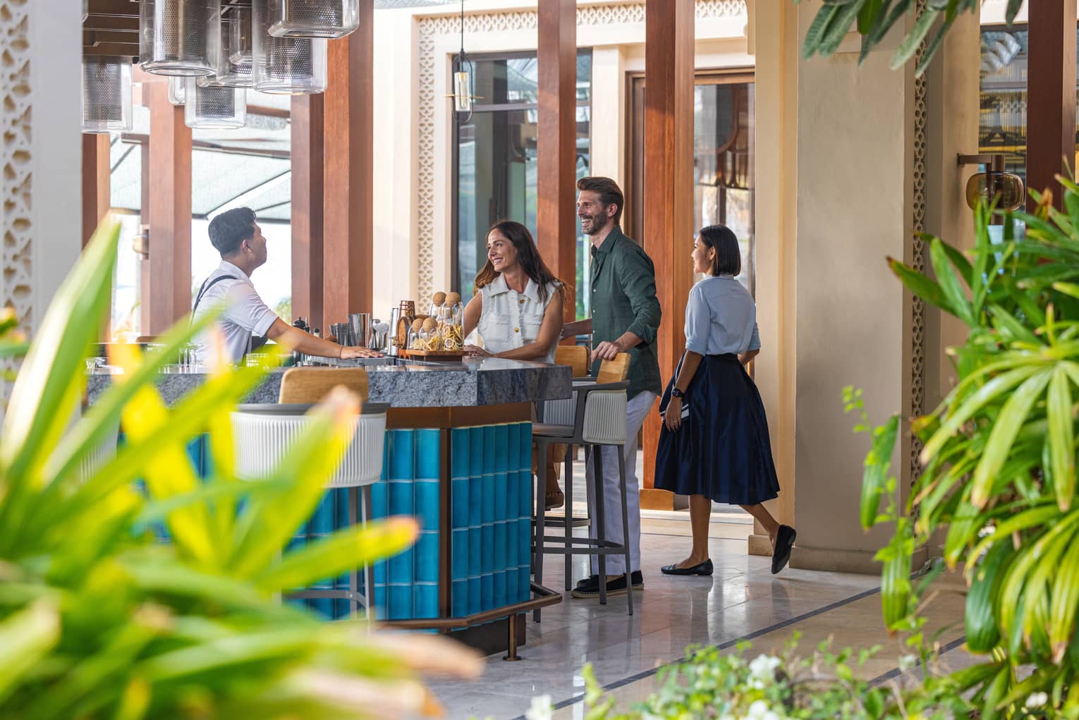 Three people walk up to an open-air, blue-tiled bar surrounded by green plants