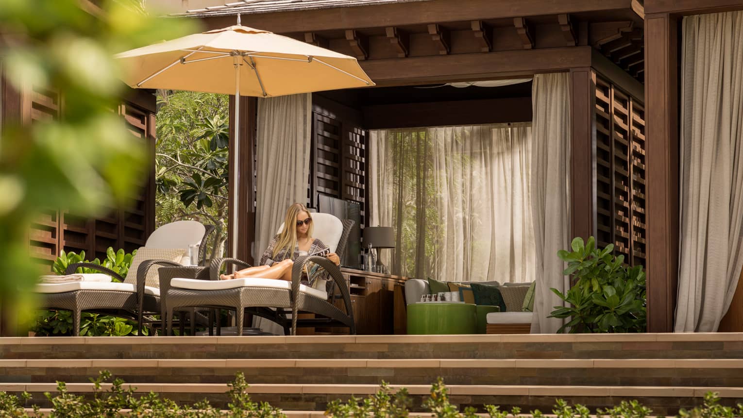 Woman reads magazine on lounge chair under white patio umbrella and poolside cabana 