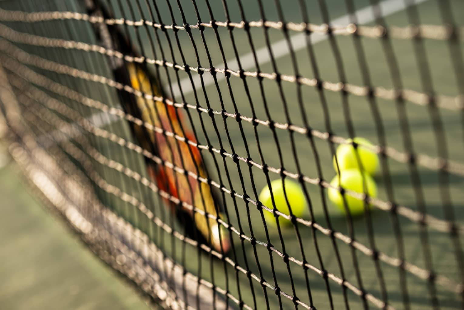 Close-up of tennis racket against net, three green tennis balls on ground