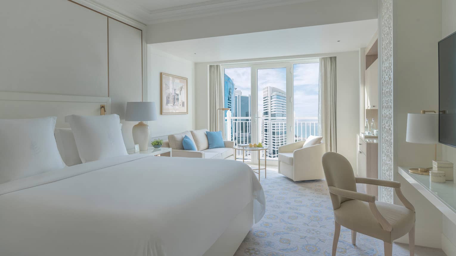 Bedroom with white bed, white walls and a white sofa and arm chair, with balcony offering city view