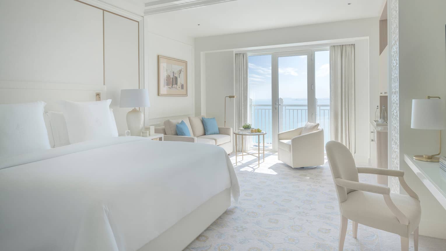 Bedroom with white bed, white walls and a white sofa and arm chair, with balcony offering sea view