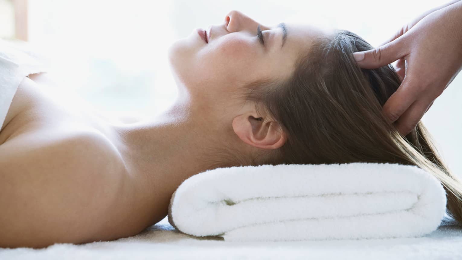Woman lays on back with head on folded white towel, closes eyes as hands massage her scalp