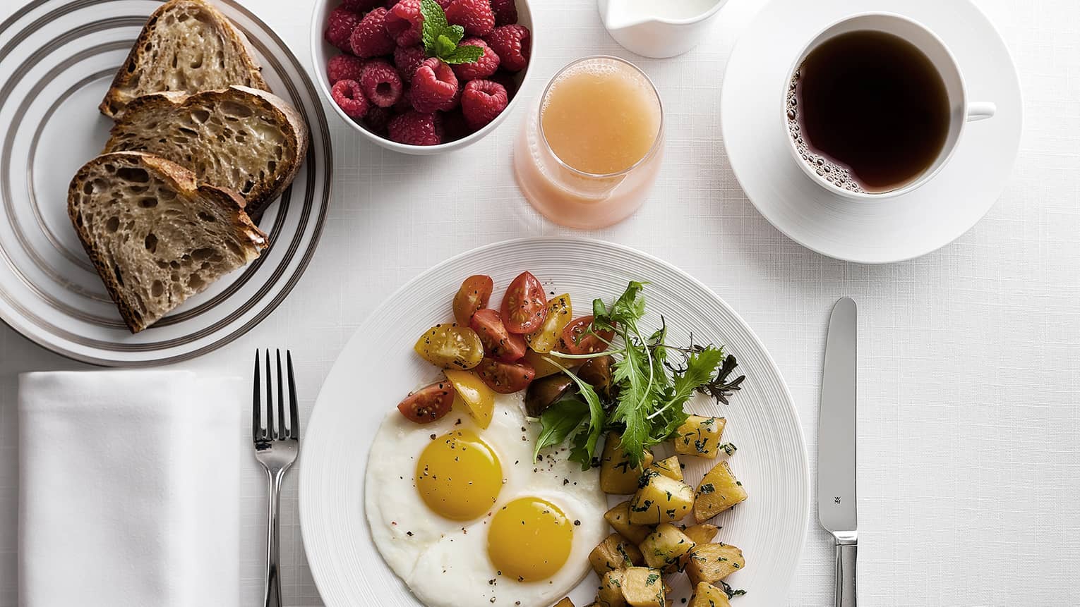 Breakfast spread with fried eggs, roasted potatoes, mixed greens, cherry tomatoes, sliced bread, a bowl of raspberries, a cup of coffee, and a glass of juice.