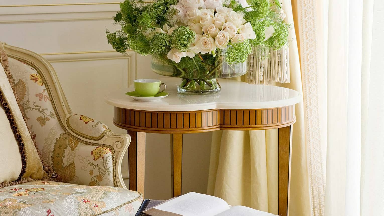 Close-up of elegant antique-style floral armchair with open books on ottoman, heart-shaped wood side table with flowers