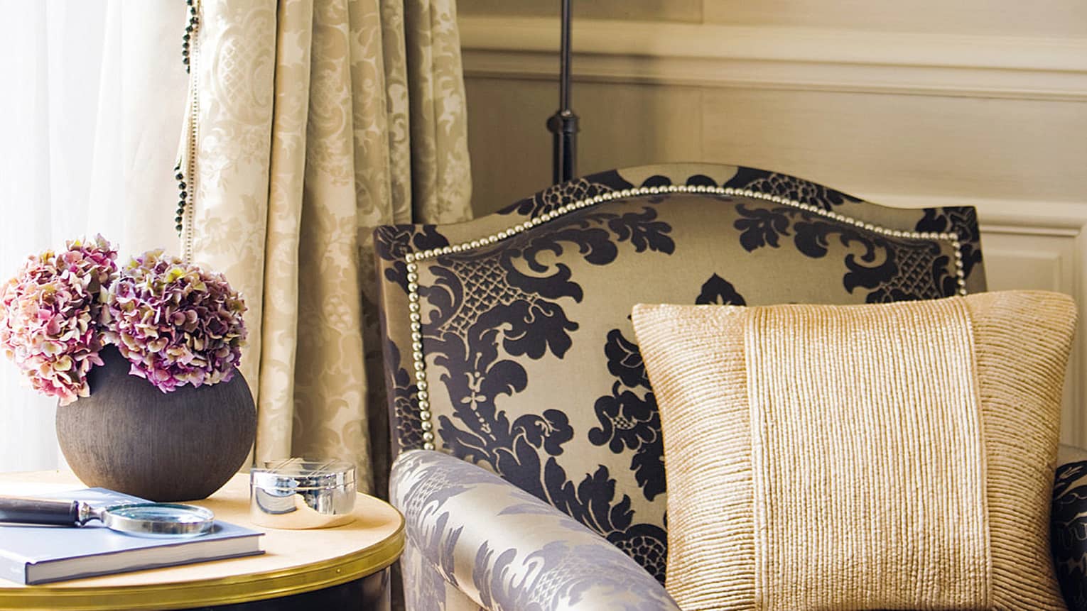 Close-up of armchair with black-and-cream Damask print, gold accent pillow, table with fresh purple flowers in black vase