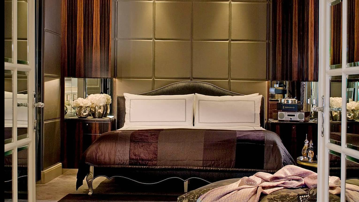 Dimly-lit hotel room, bed with deep brown and purple satin bedspread, tall fabric headboard and wood panel walls