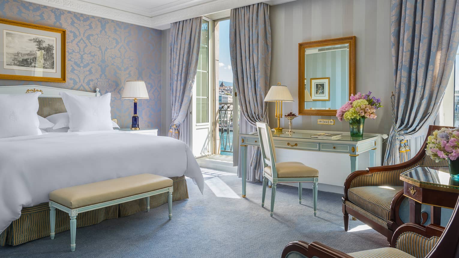 Elegant hotel room with a king bed, desk, seating area and a balcony overlooking water, featuring light blue and gold decor