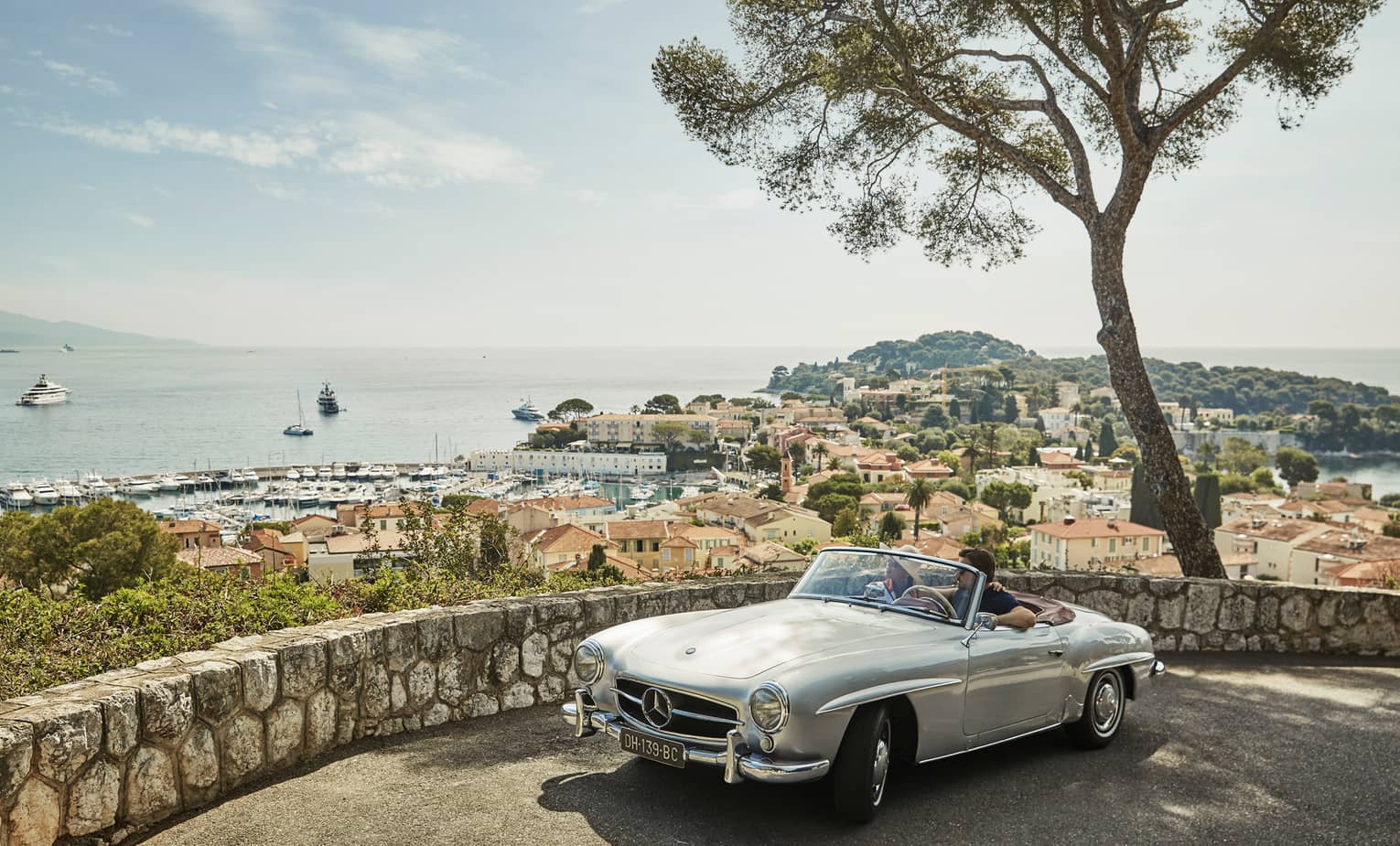 Couple in luxury vintage white car parked under tree by stone wall overlooking red roofs, water