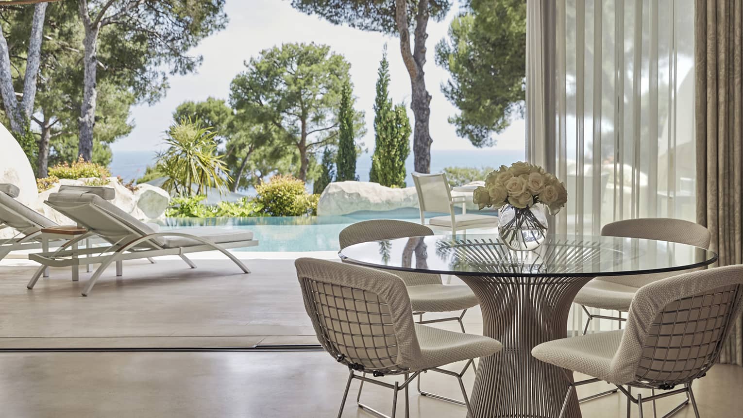 Hotel suite with round glass table and chairs with view of lounge chairs on patio and private pool surrounded by trees