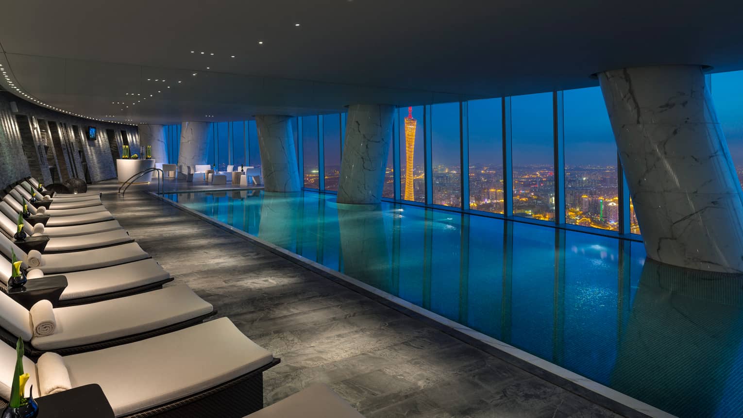 White chairs and indoor pool on 69th floor at night, beside windows overlooking city lights and Pearl River