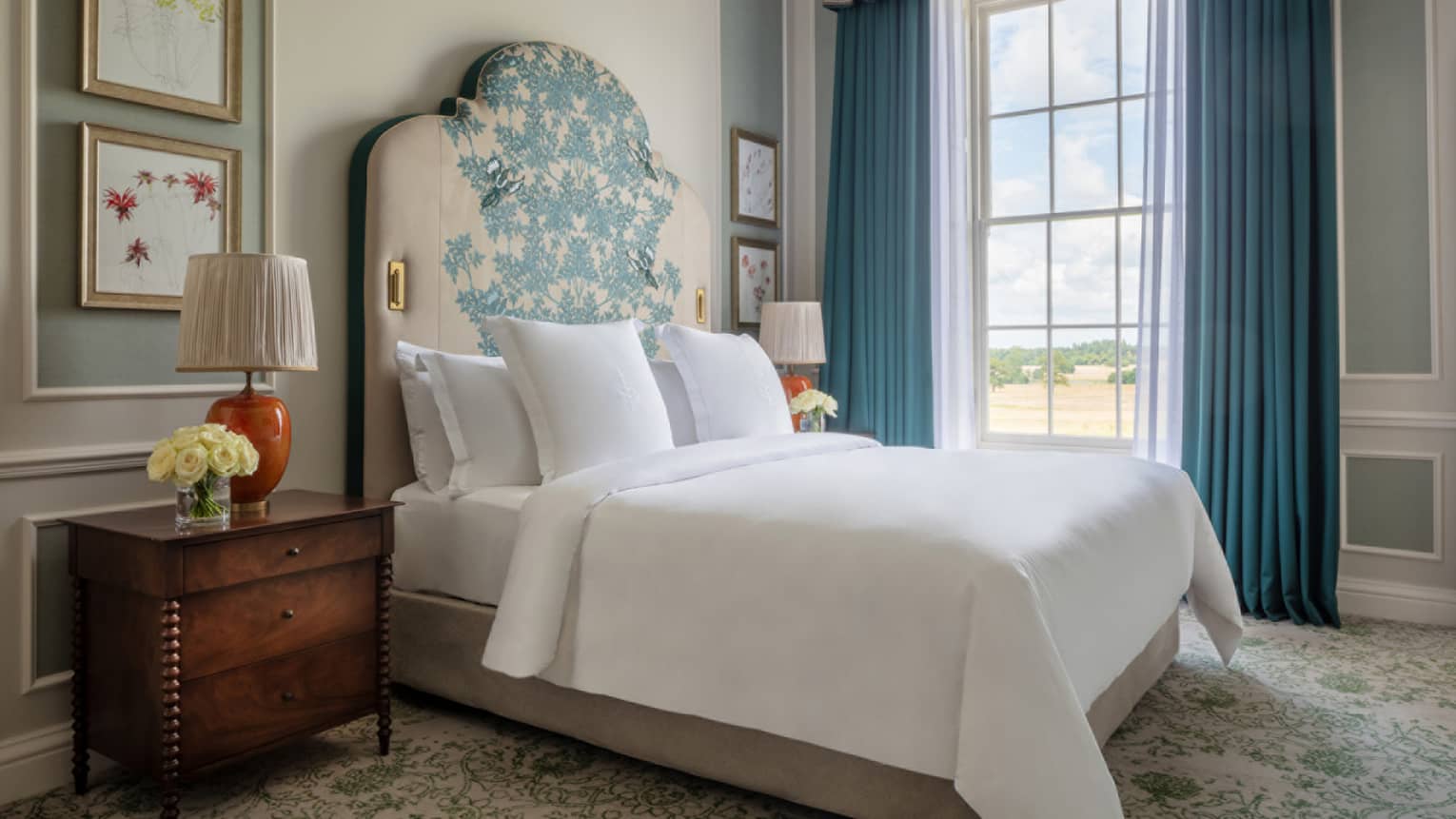 Guest room with Queen bed, white linens, ornate headboard, paisley carpeting, cherry bed side table, window with blue drapes