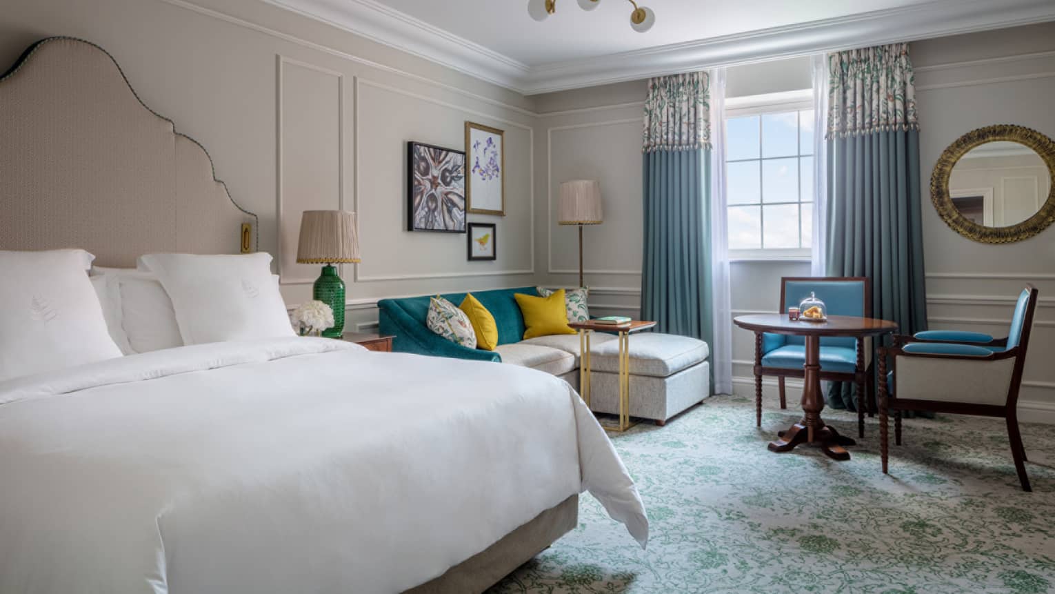 Guest room with king bed and white linens, cream sectional sofa with colourful pillows, small round table and two chairs, window with blue drapes