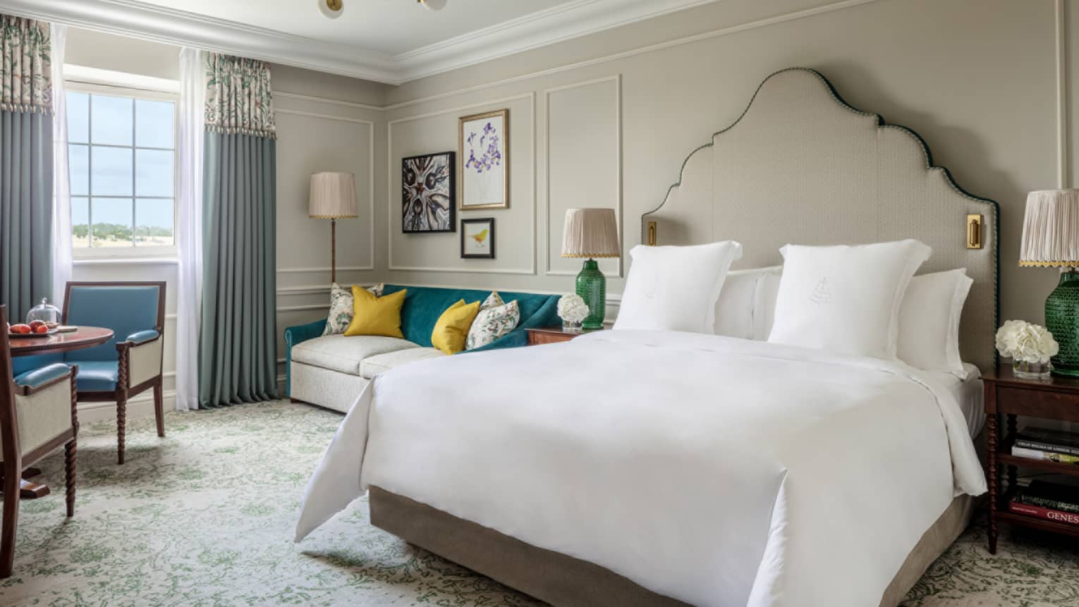 Guest room with king bed and white linens, cream couch with colourful pillows, small round table and two chairs, window with blue drapes