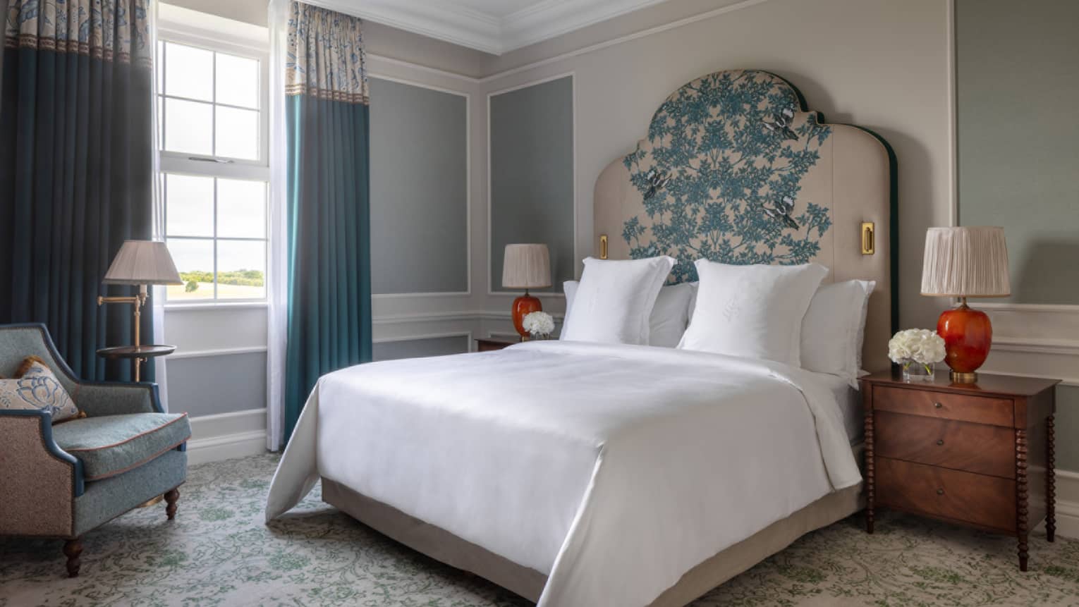 Guest room with Queen bed and white linens, embroidered headboard, cherry bed side tables with red lamps, turquoise accent chair, window with blue drapes