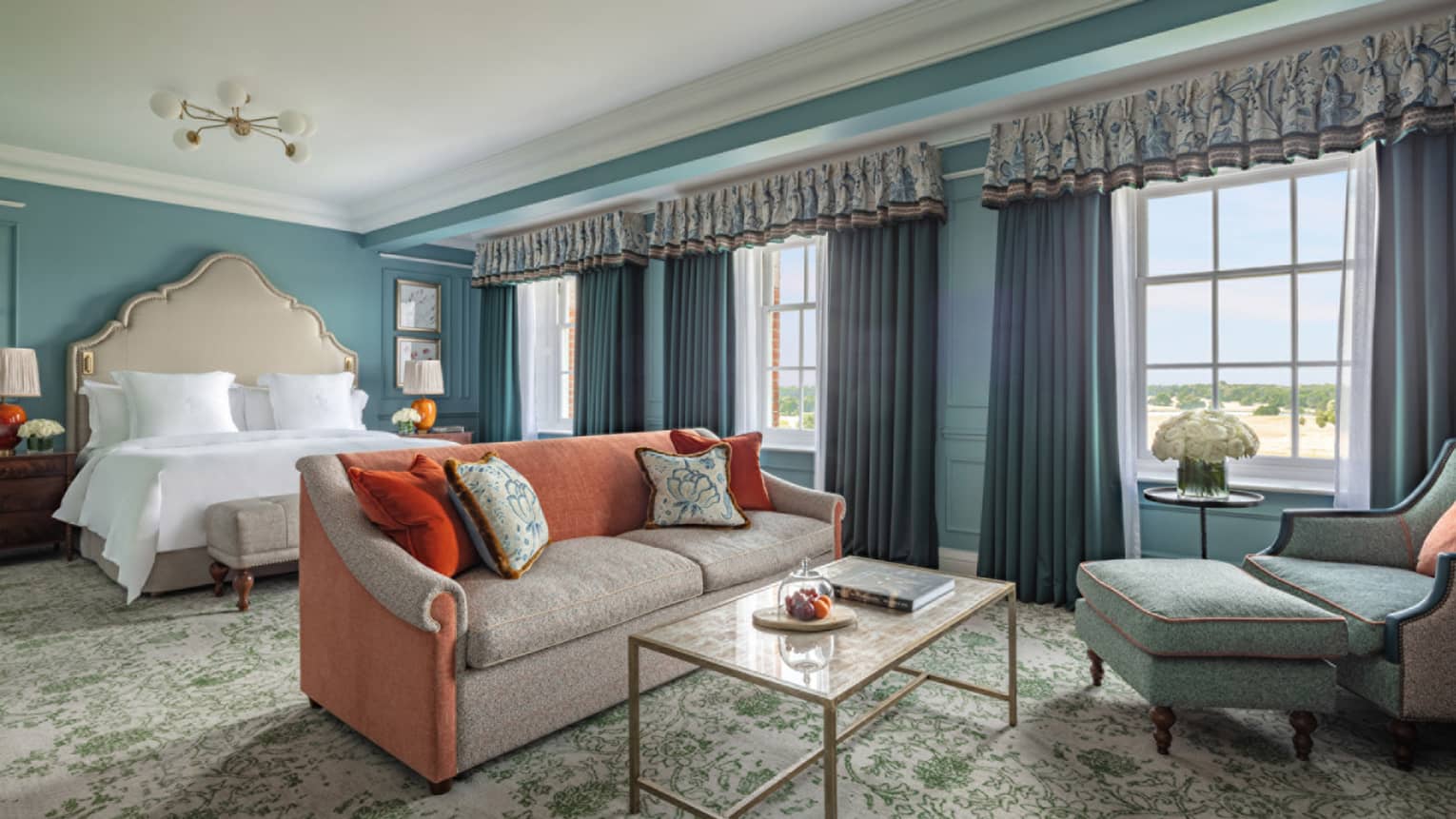 Guest room with Queen bed, cream and red couch, coffee table, blue accent chair, trio of windows with blue drapes