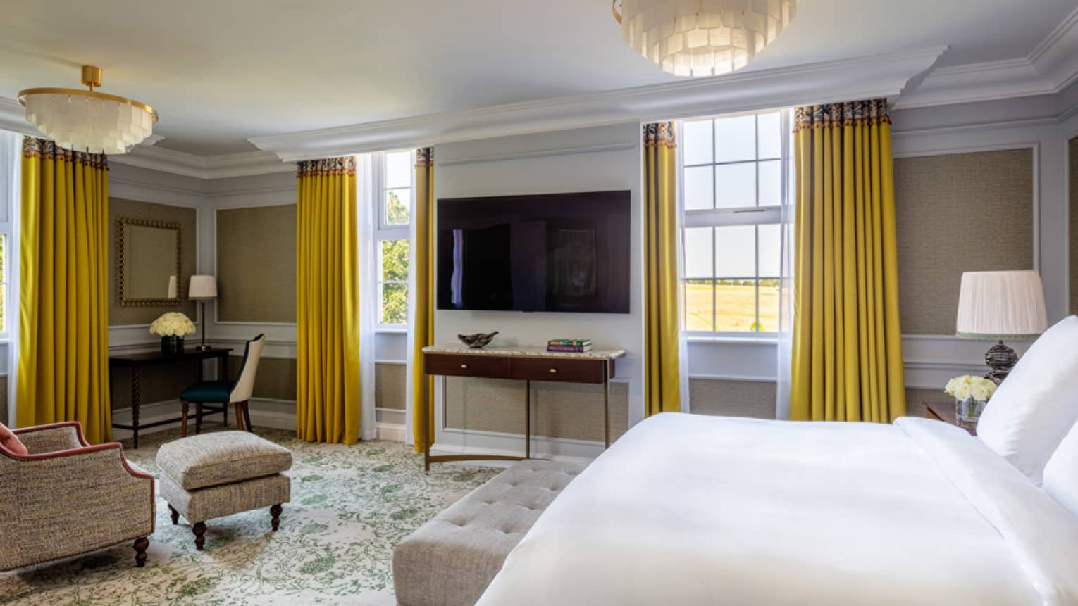 Guest room with Queen bed, cream-coloured accent chair, dark wood desk and chair in corner, trio of windows with yellow drapes