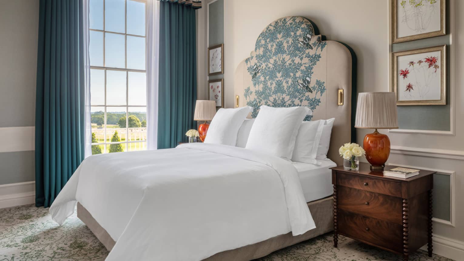 Guest room with Queen bed with white linens and embroidered headboard, cherry bedside table with orange lamp, floor-to-ceiling window with blue drapes