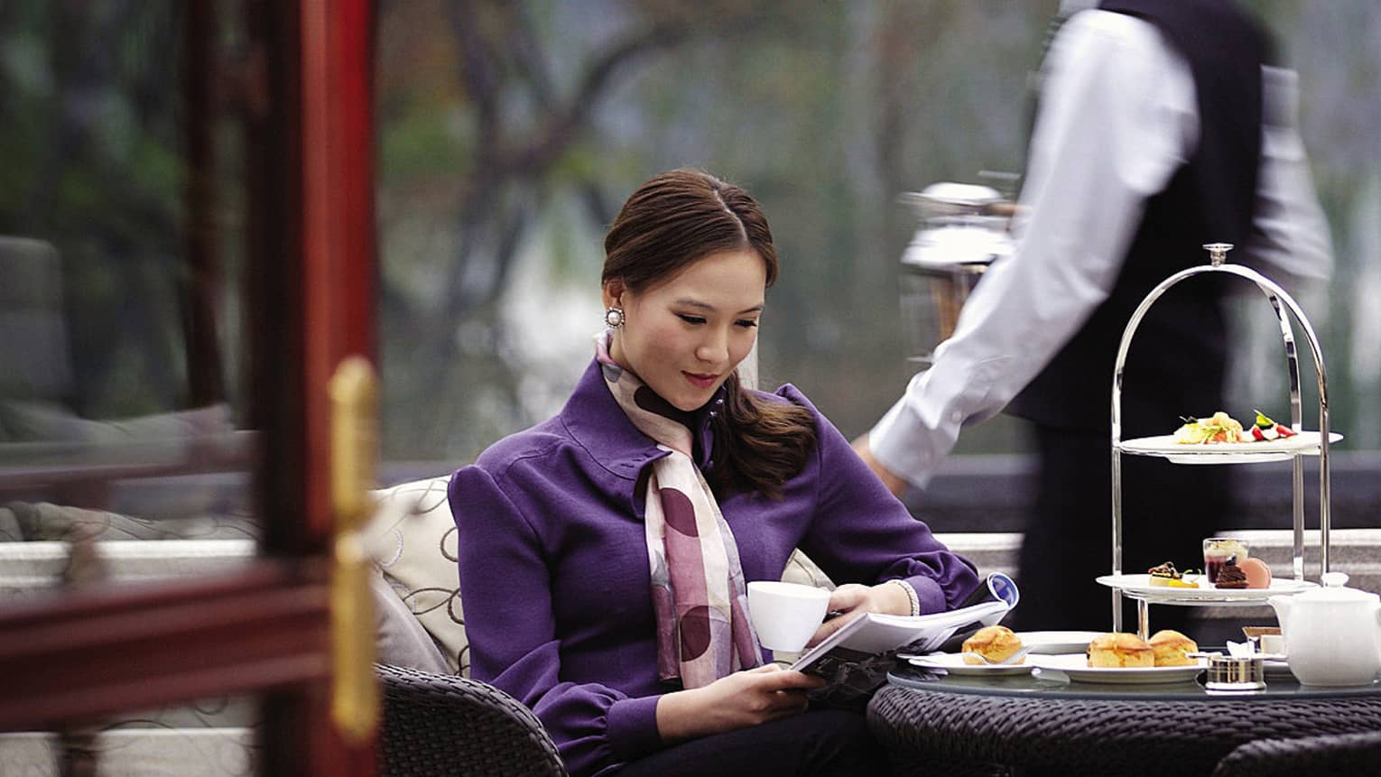 Woman wearing purple coat, seated at afternoon tea in Lobby Lounge, staff member behind