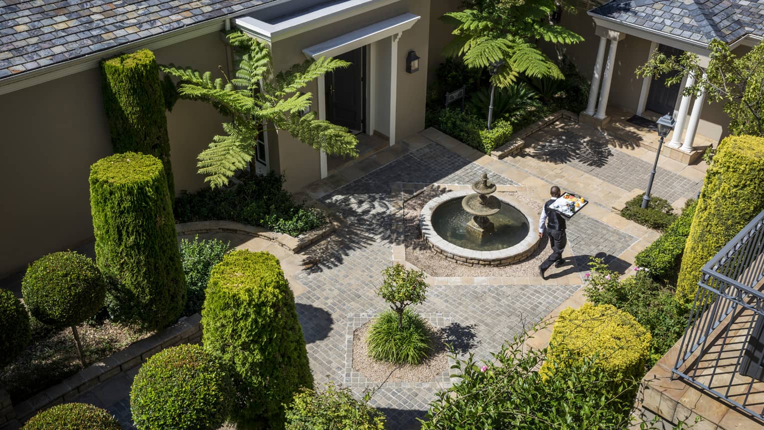 Aerial view of waiter carrying tray through brick courtyard with manicured green shrubs
