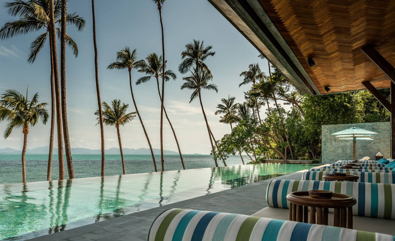 Green-and-blue striped banquettes on patio by infinity swimming pool, ocean