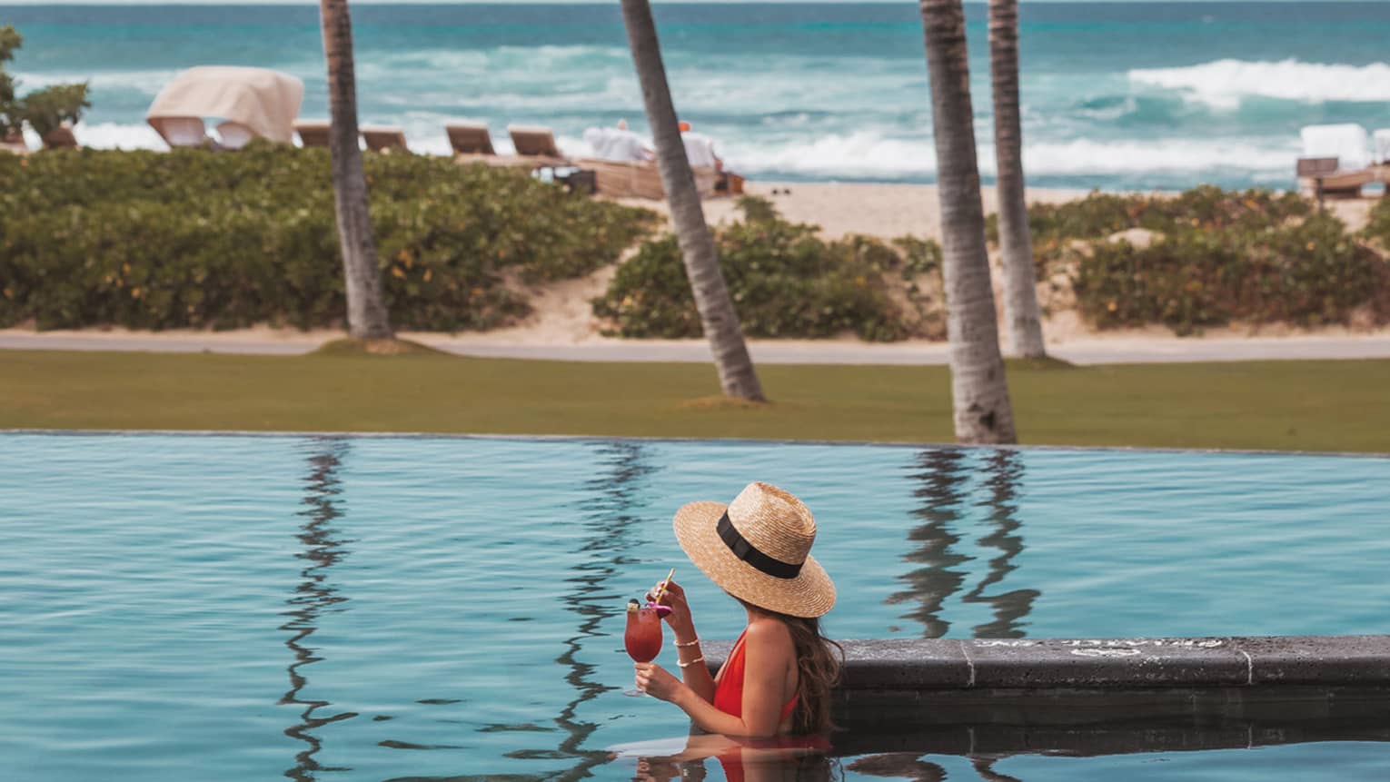 Side view of person in pool wearing straw sunhat and holding drink, ocean view