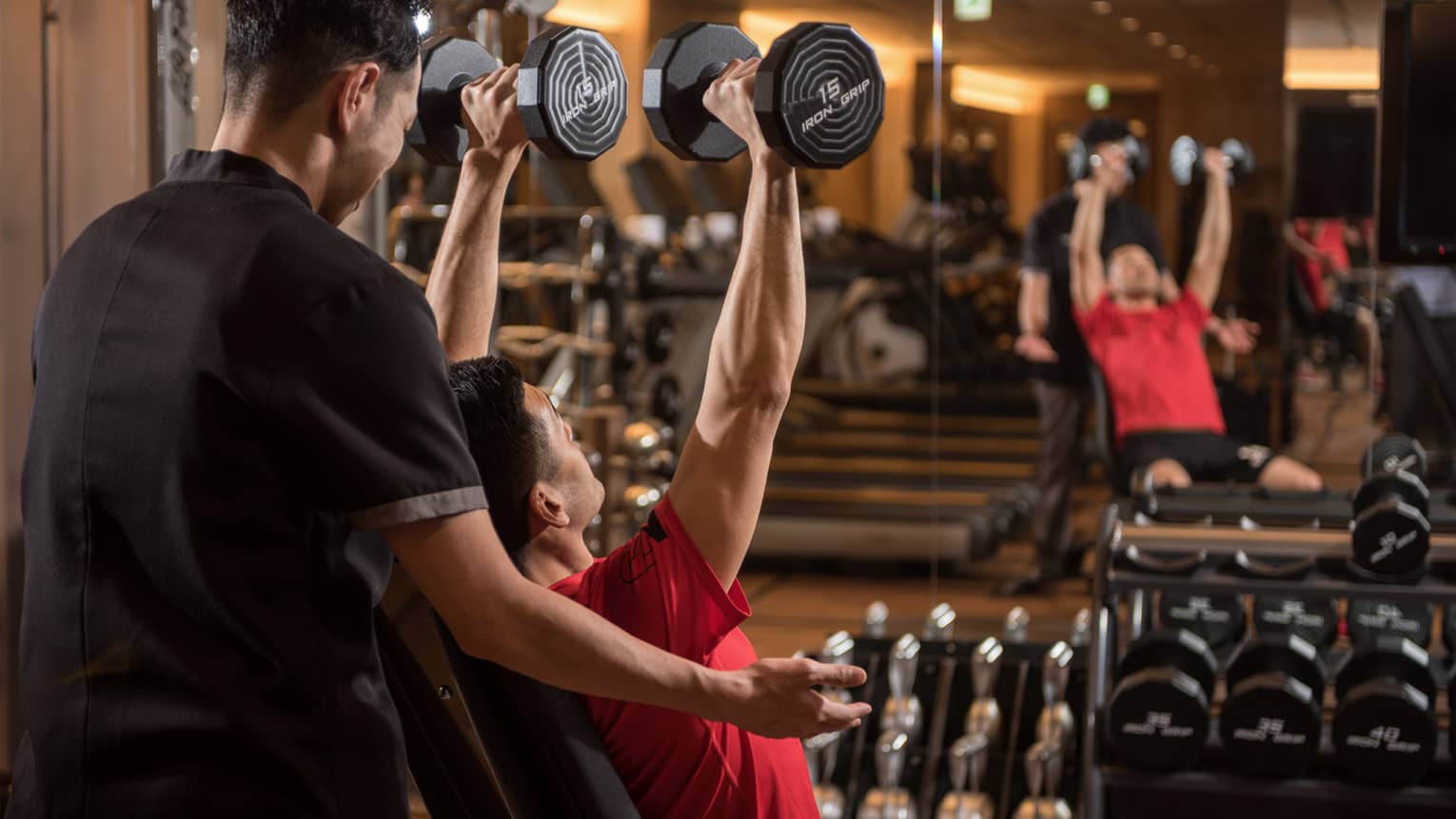 Man in chair in front of mirror lifts two 15-pound weights above his head while personal trainer assists
