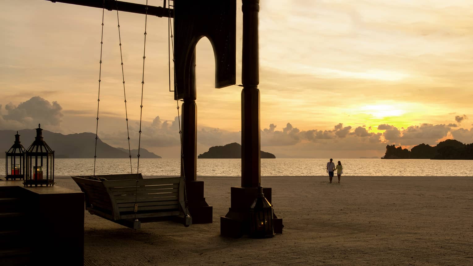 Silhouettes of couple walking along beach past wood swing, pavillion at sunset