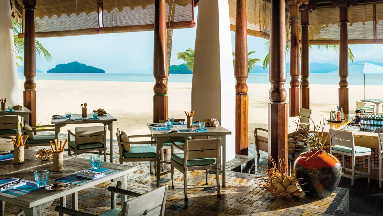 Dining tables, chairs under tall carved wood pillars with open-air walls to beach