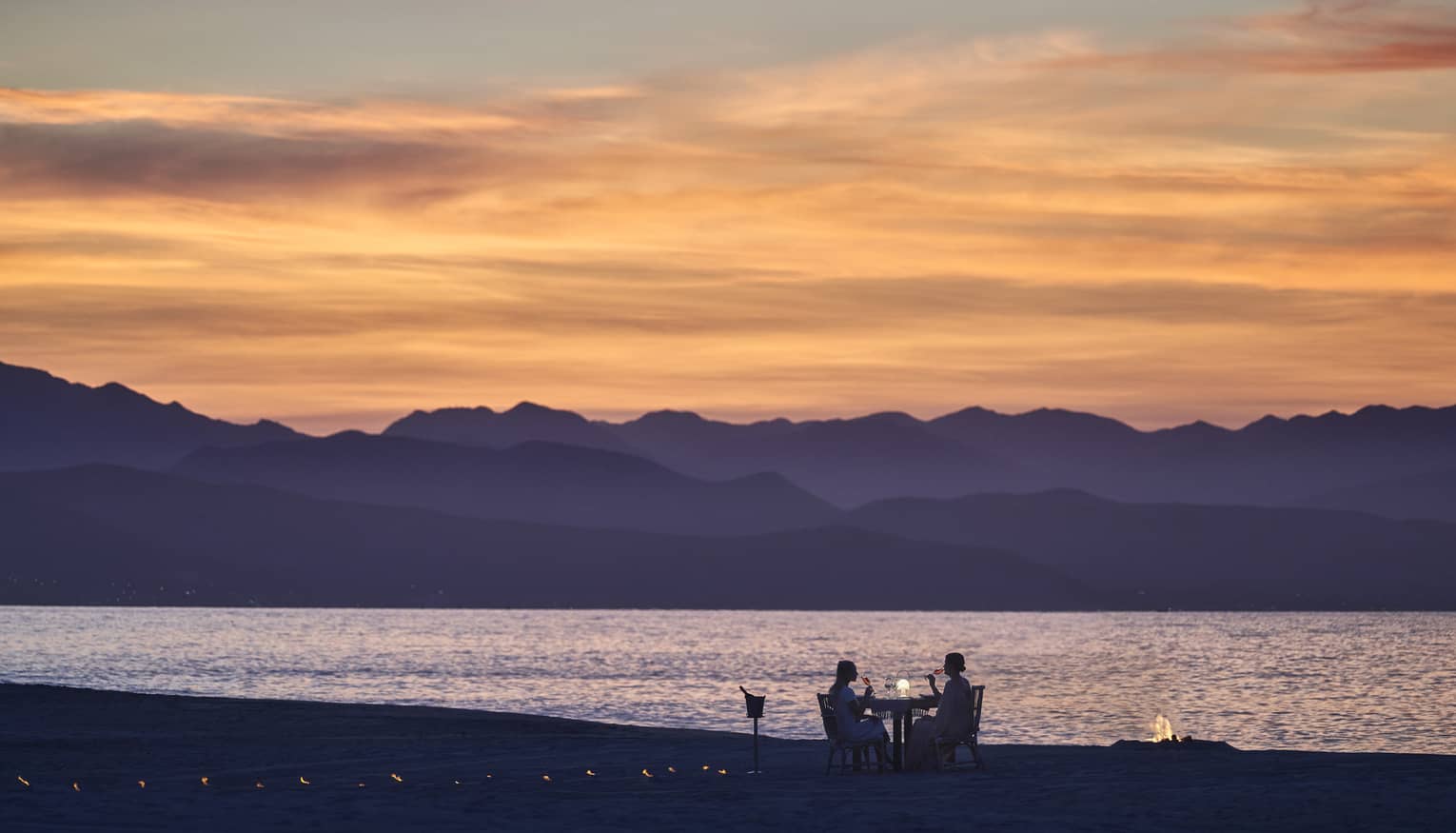 A couple dining on a beach during sunset, the mountains in the background look purple while the sky is orange and pink.