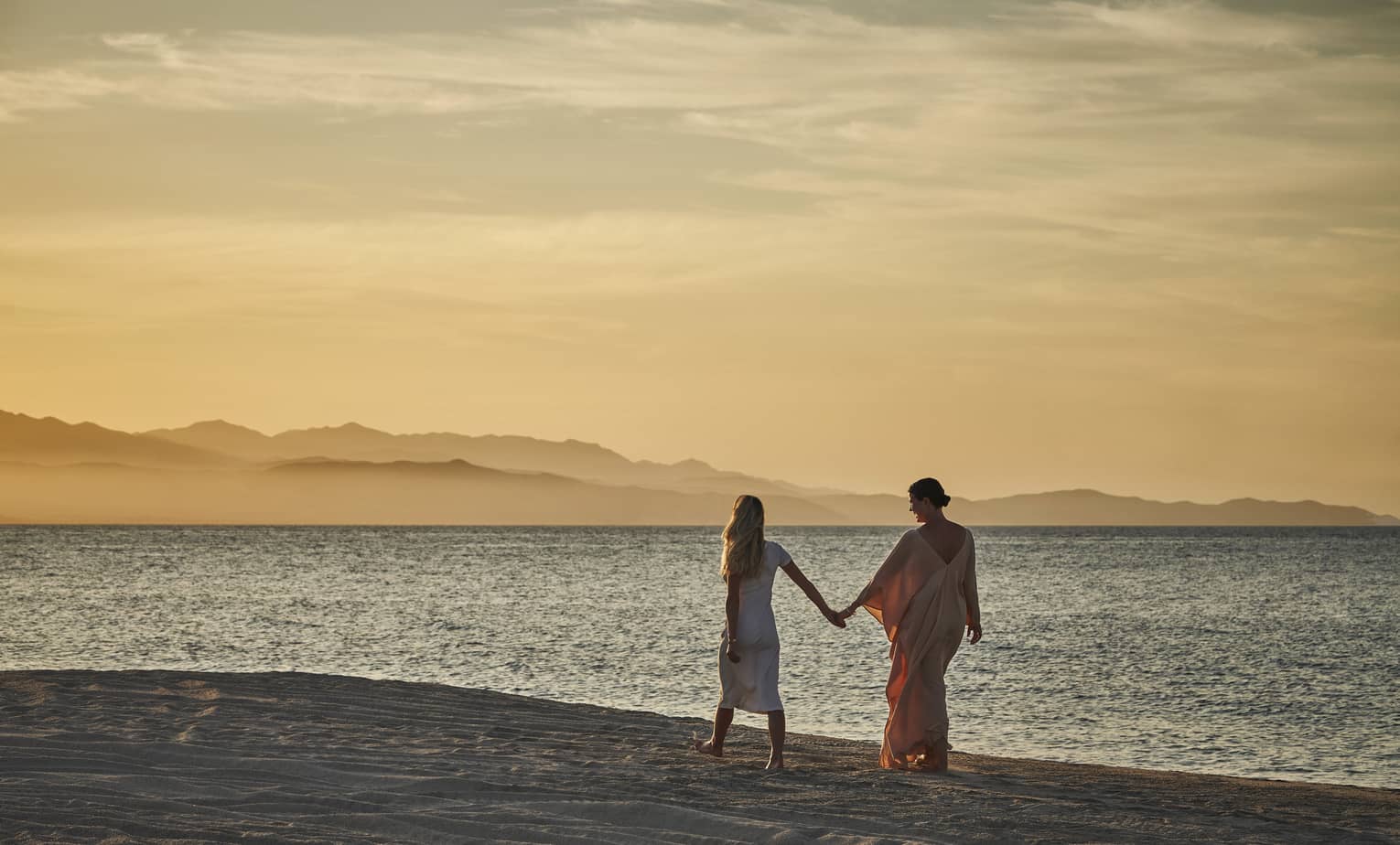 Two women holding hands and walking along a beach while the sun is setting.