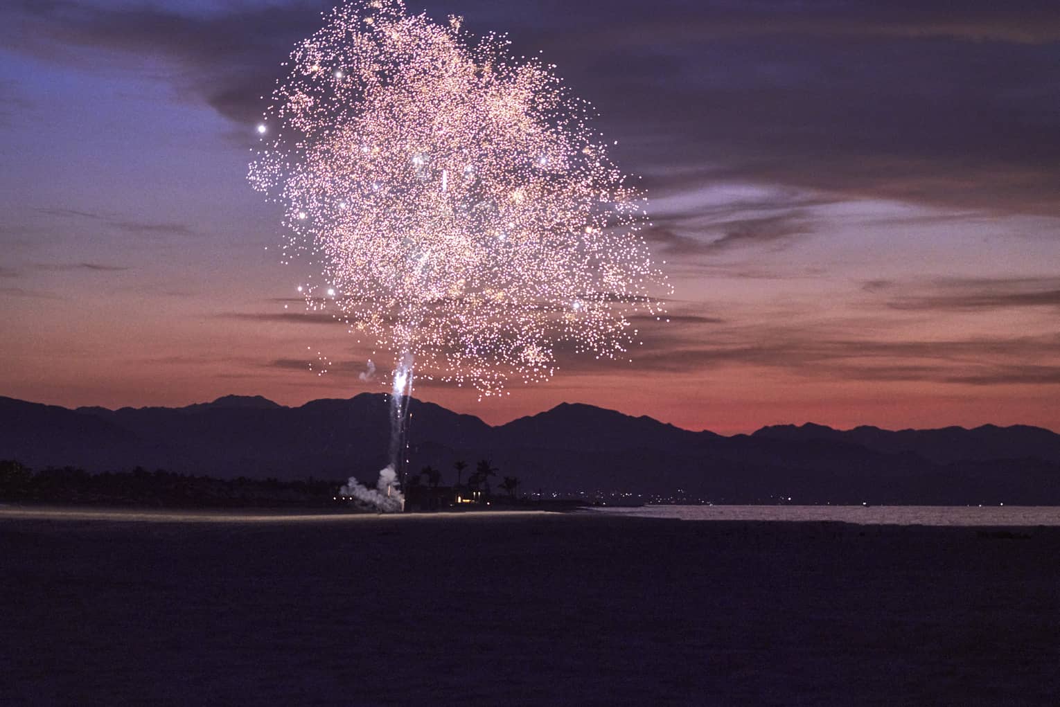 Pink fireworks exploding during sunset with a mountain behind them.