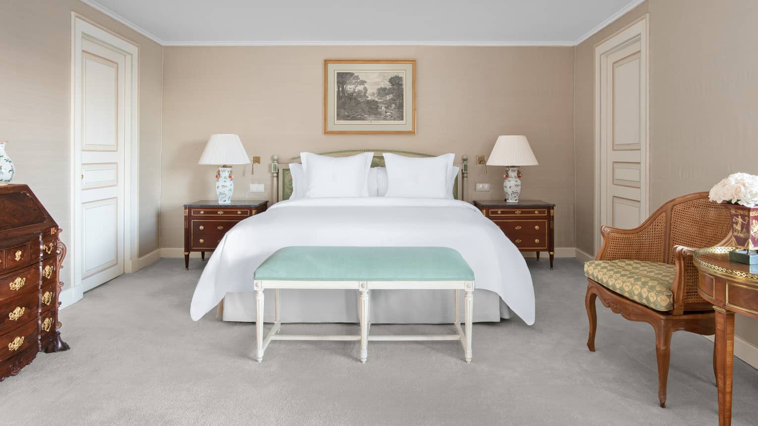 Beige-walled guest room with white bed flanked by wooden nightstands and white lamps, blue foot bench