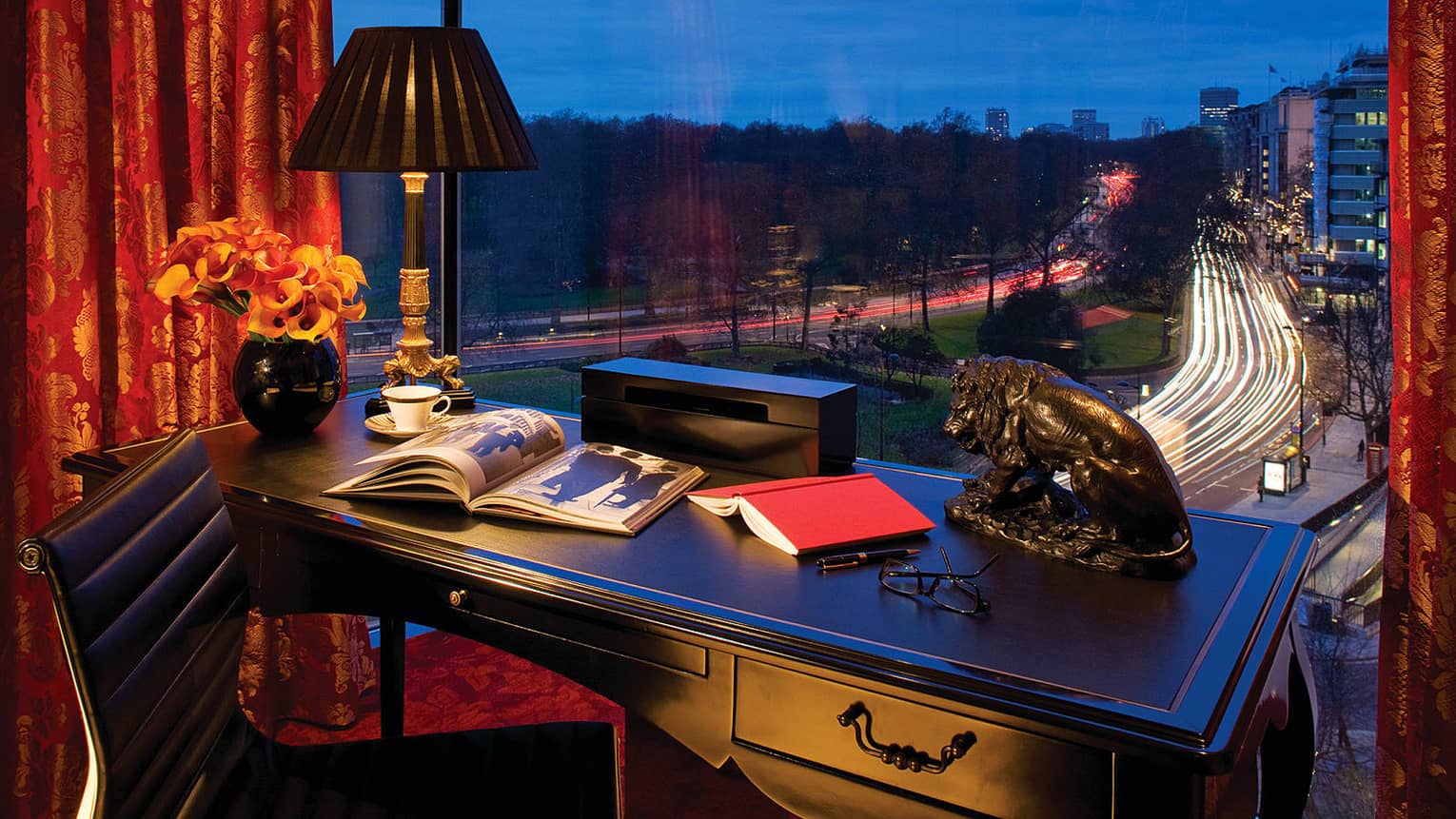 Window-facing black wooden desk topped with lion figurine and books, black leather chair, view of the bustling city at night
