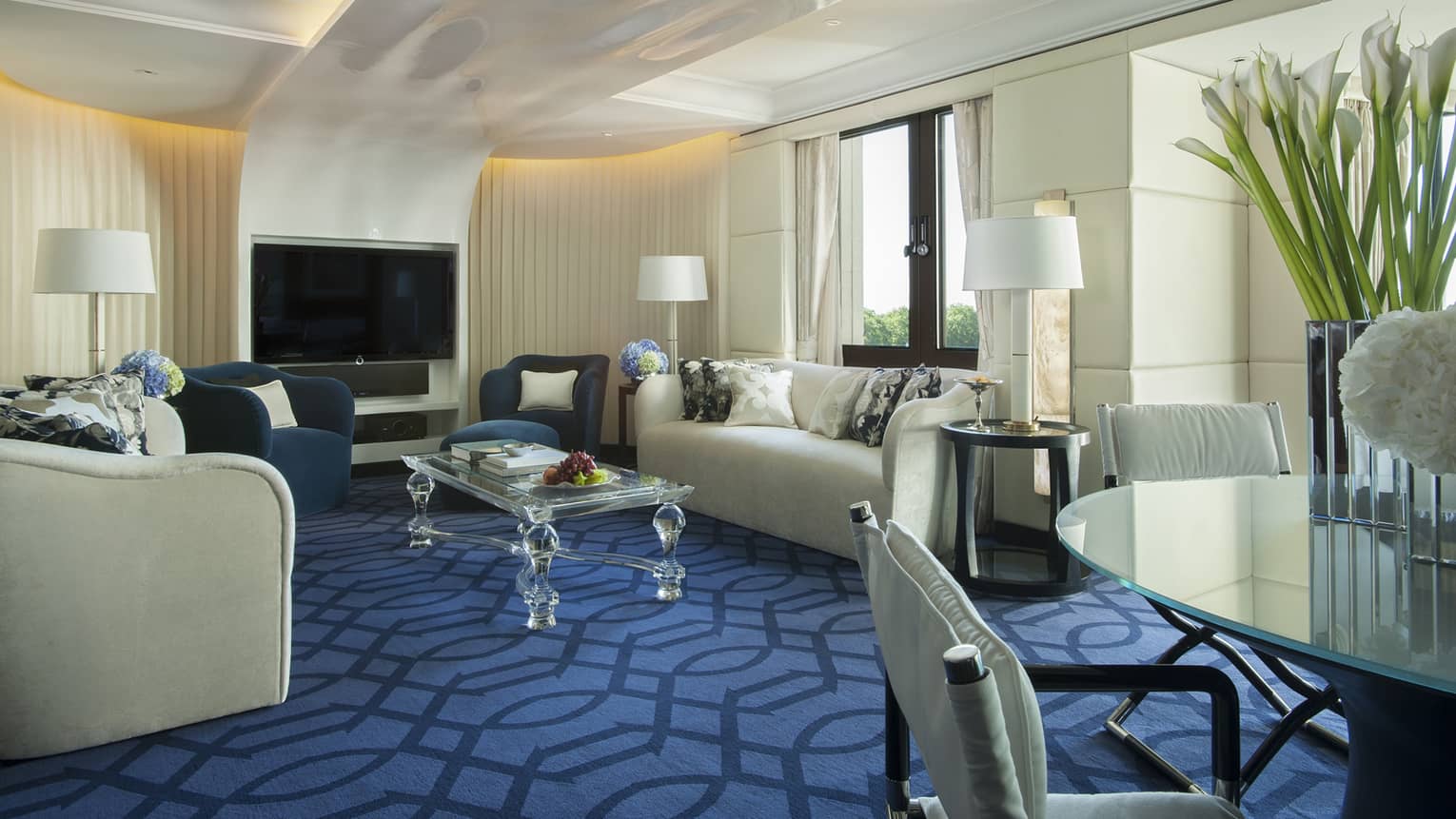 The Blue Suite at Four Seasons London Park Lane in Mayfair features an opulent living area with a vibrant blue and cream color scheme. The room includes two plush blue armchairs and a large cream sofa, complemented by stylish throw pillows. A modern glass coffee table holds decorative items and books, adding a personal touch. Large floor lamps and a sleek flat-screen TV are also part of the decor. Floor-to-ceiling windows provide ample natural light and a view of the green surroundings. The room's luxury is accentuated by tall cream drapes and a patterned blue carpet.