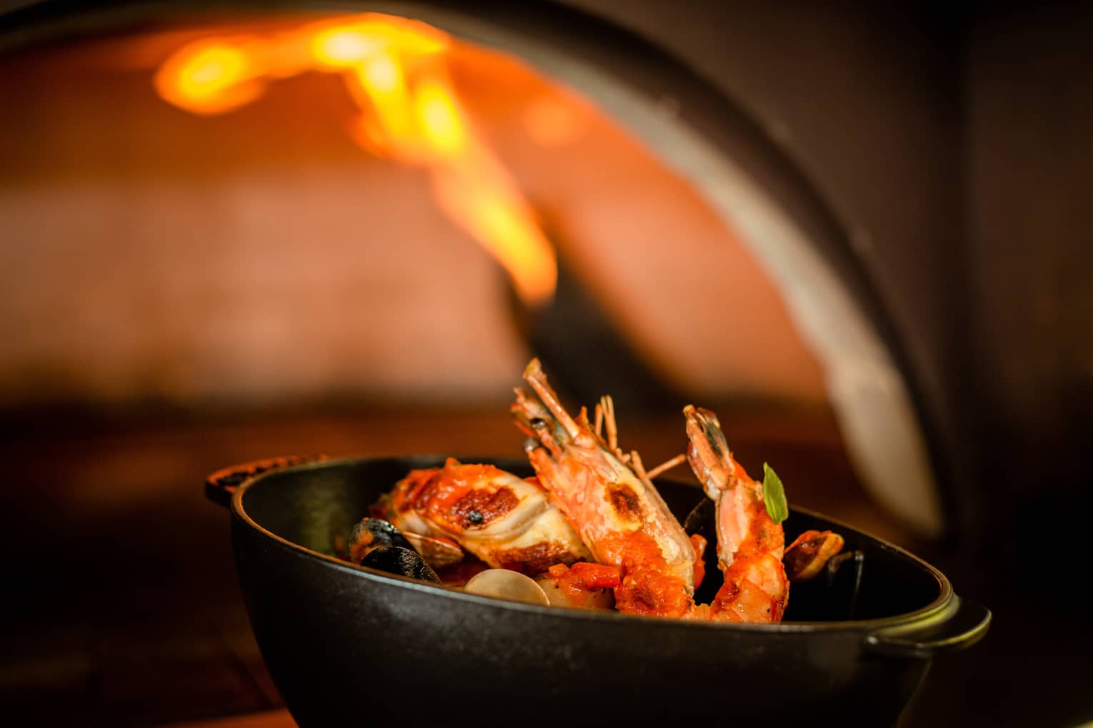 Cioppino with head-on prawns in cast iron serving dish in front of pizza oven