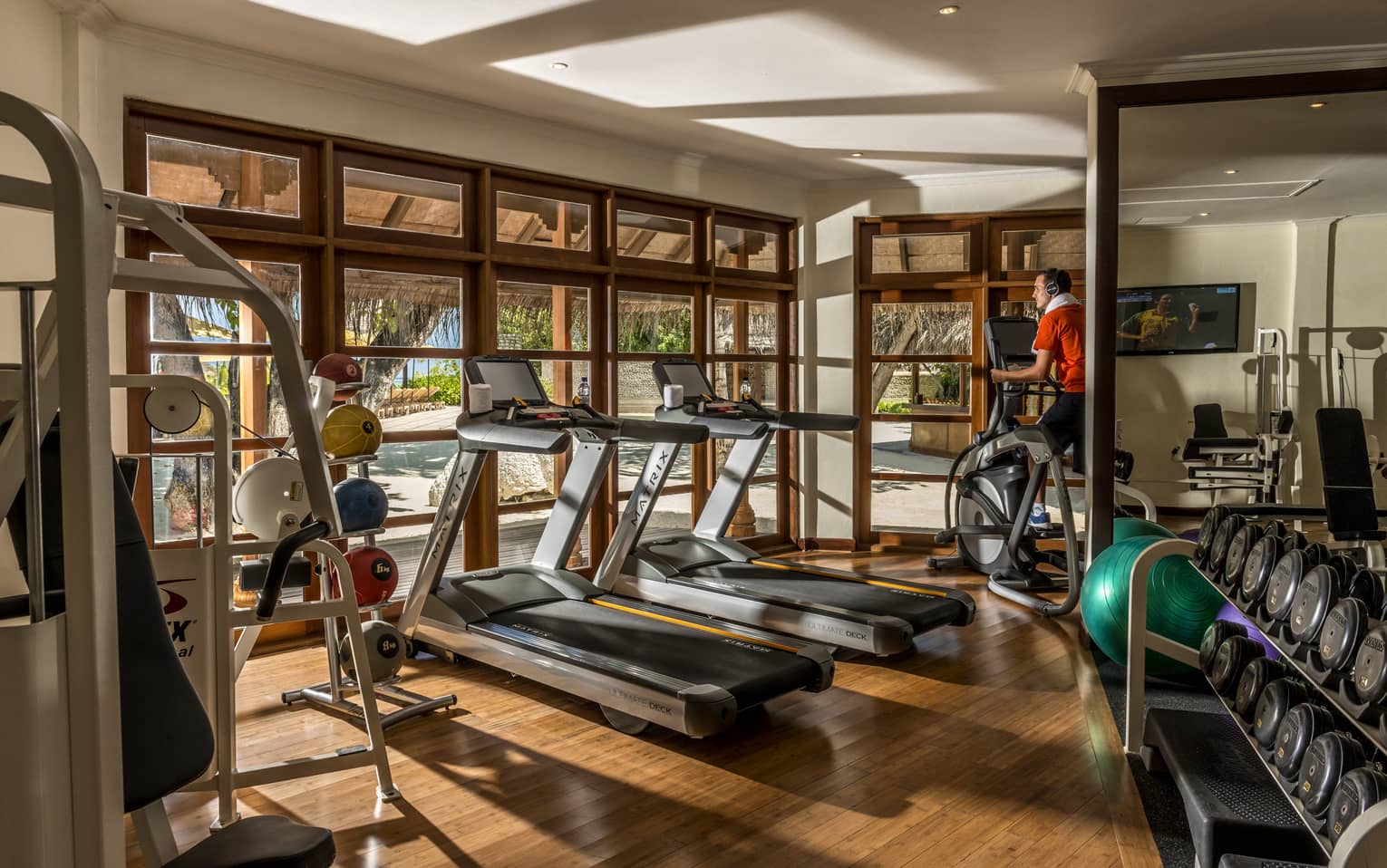 Gym with wooden floors, two treadmills, person on elliptical, free wights, windows looking out to beach