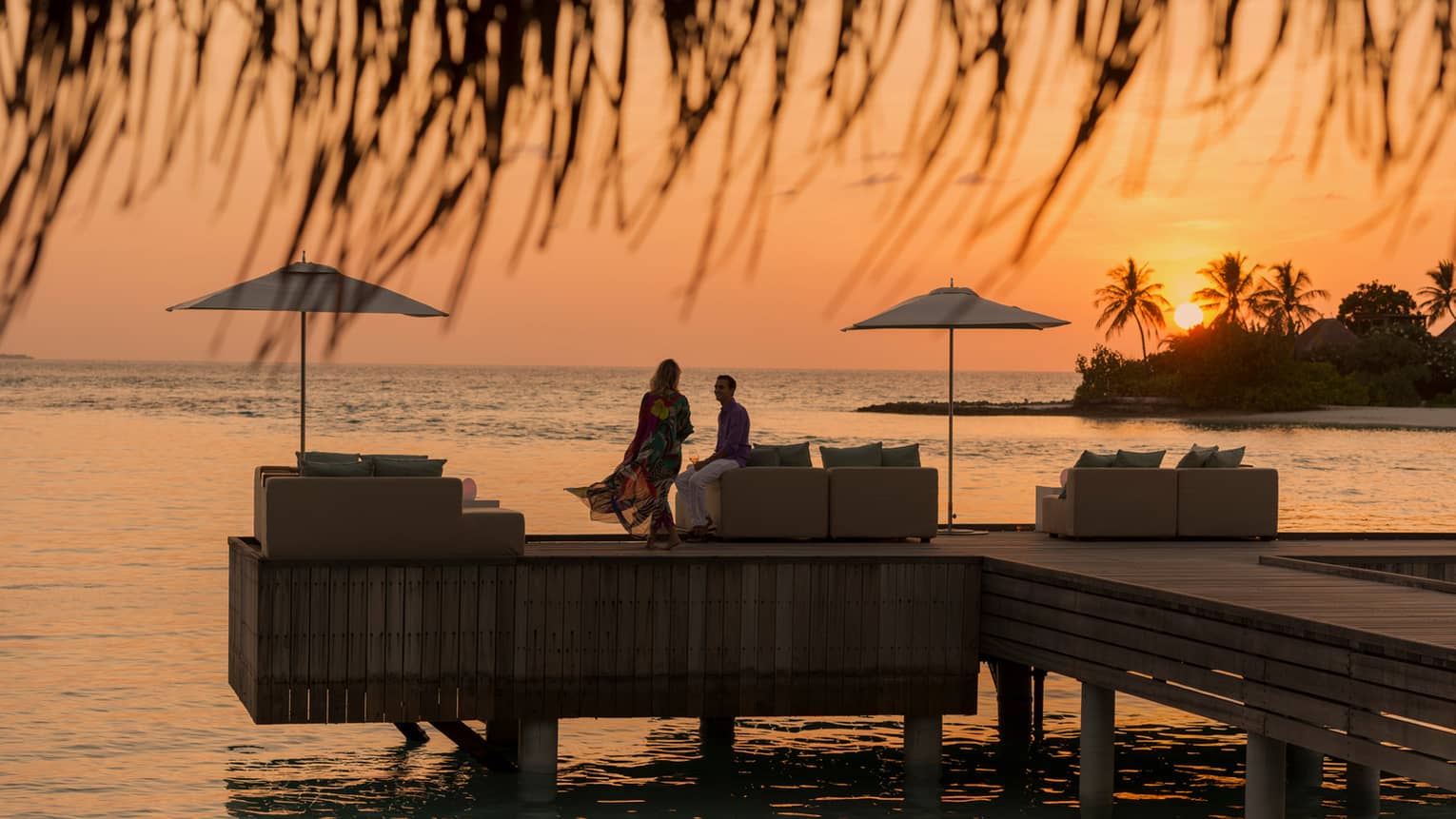 Private island dining at sunset. Woman with long dress blowing in the wind, man sits on the edge of white sofa