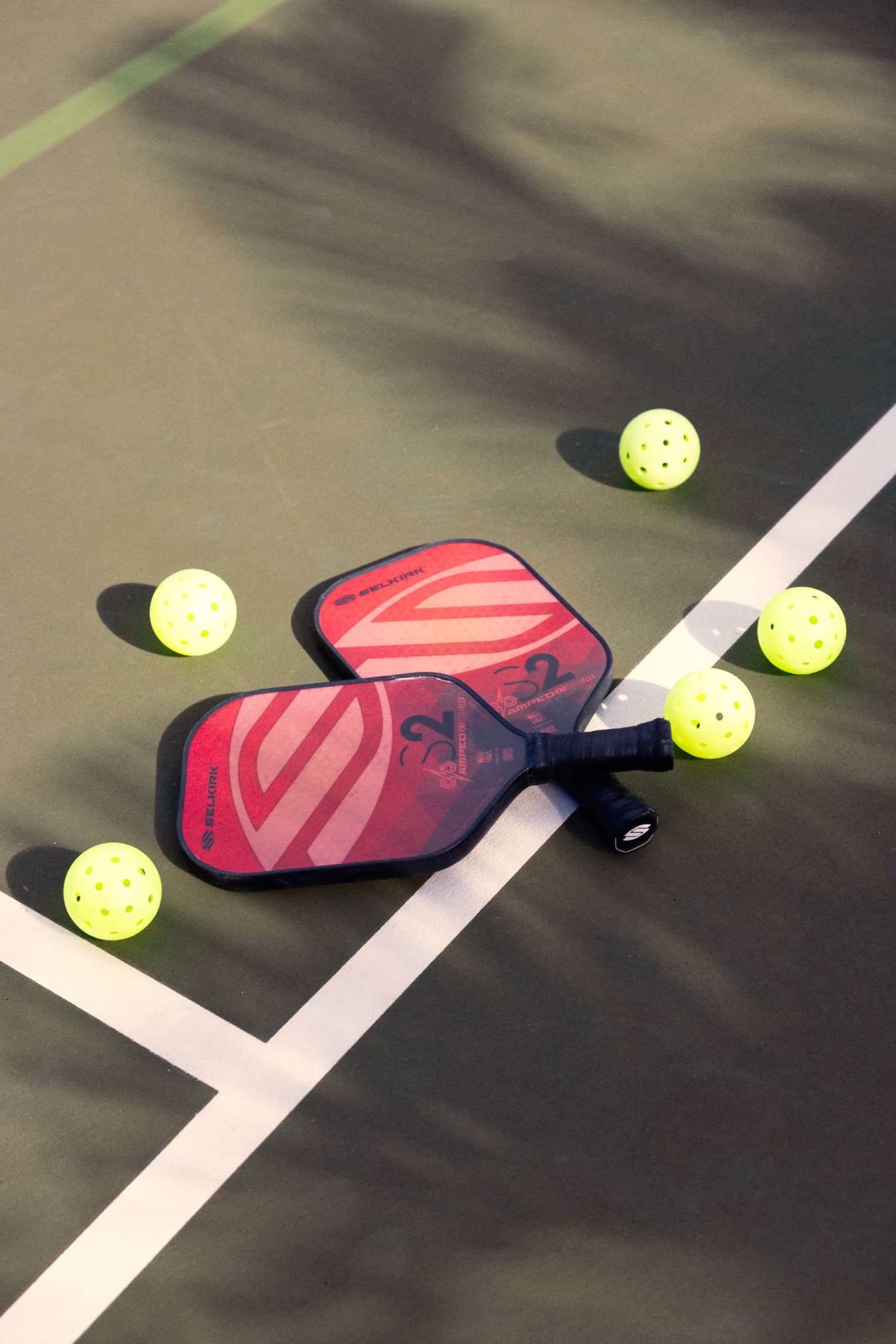 Close-up of two red and pink pickleball racquets, handles crisscrossed on a white court line, surrounded by five pickleballs.