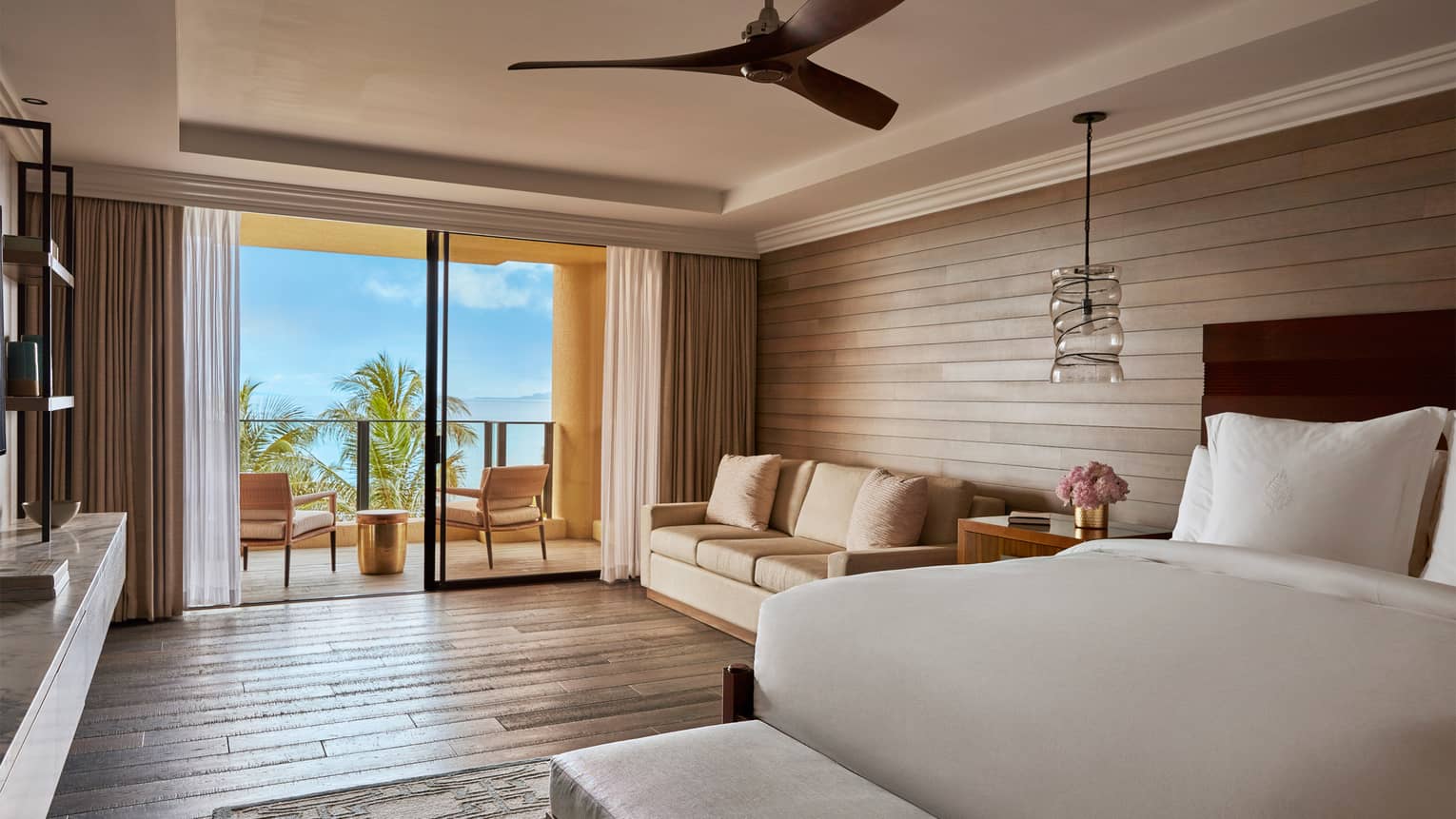 "Elite" suite bedroom with bed, hardwood floors and walls, beige loveseat, balcony with chairs, brass table