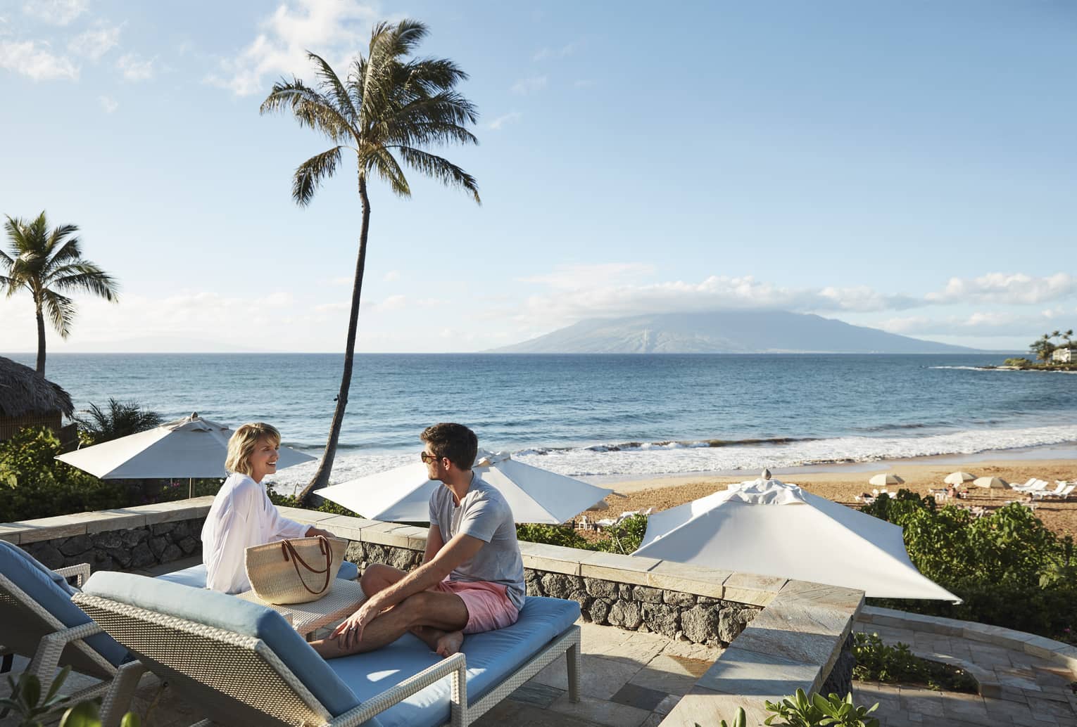 Man in shorts and smiling woman in beach shirt sit on edge of lounge chair, beach and ocean in background