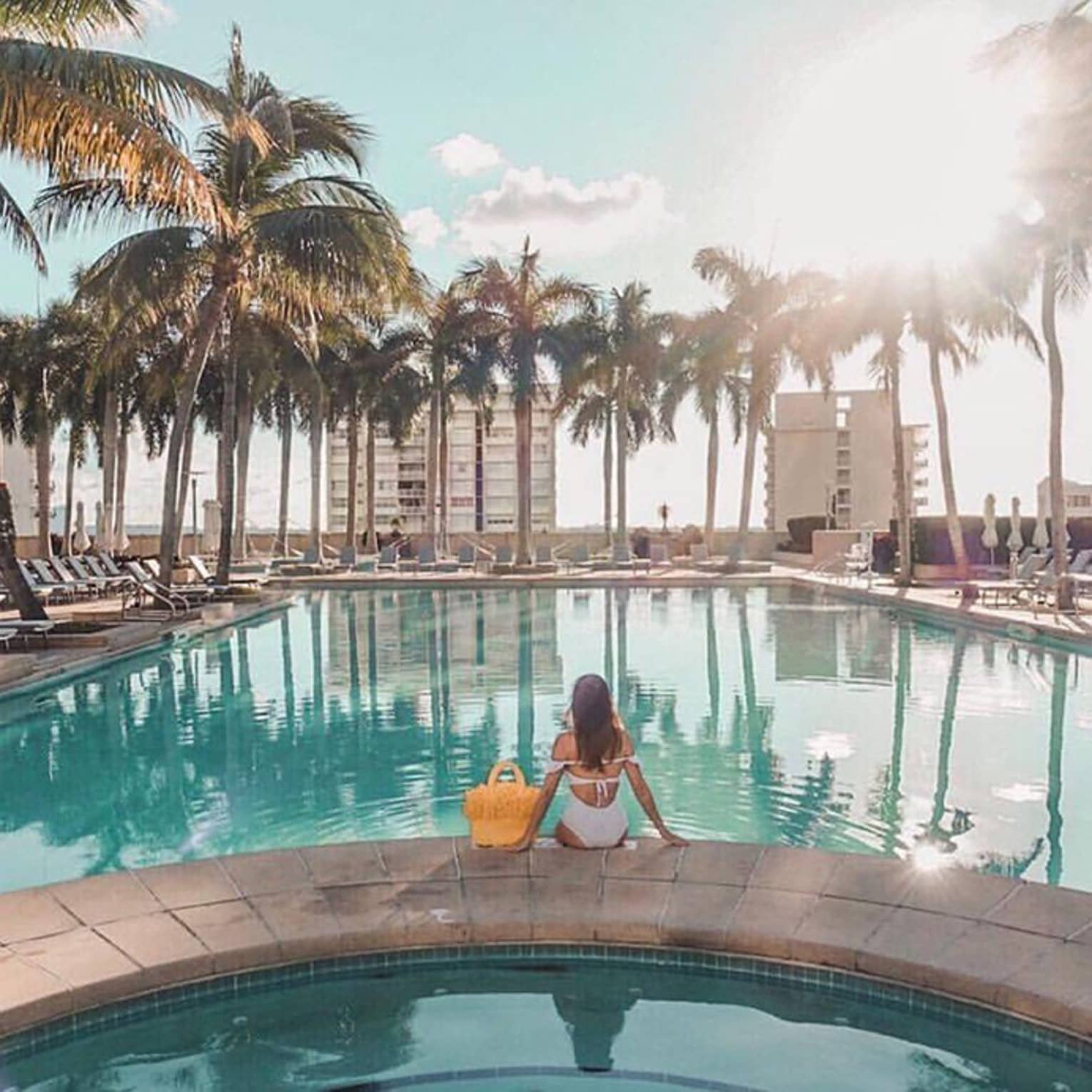 Woman sits on edge of outdoor swimming pool, facing palm trees and building