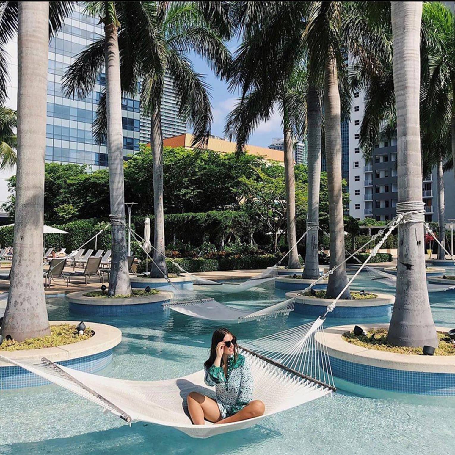 Woman lounges in hammock over outdoor swimming pool with potted palm trees