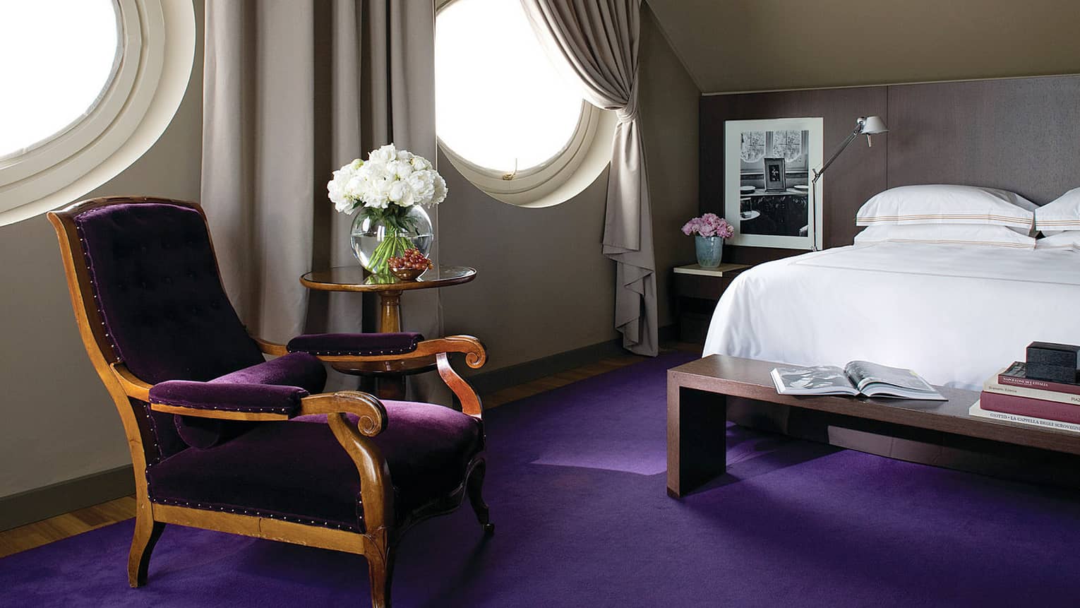 Designer Suite circular windows over purple velvet chair, carpet, bed with wood bench, books