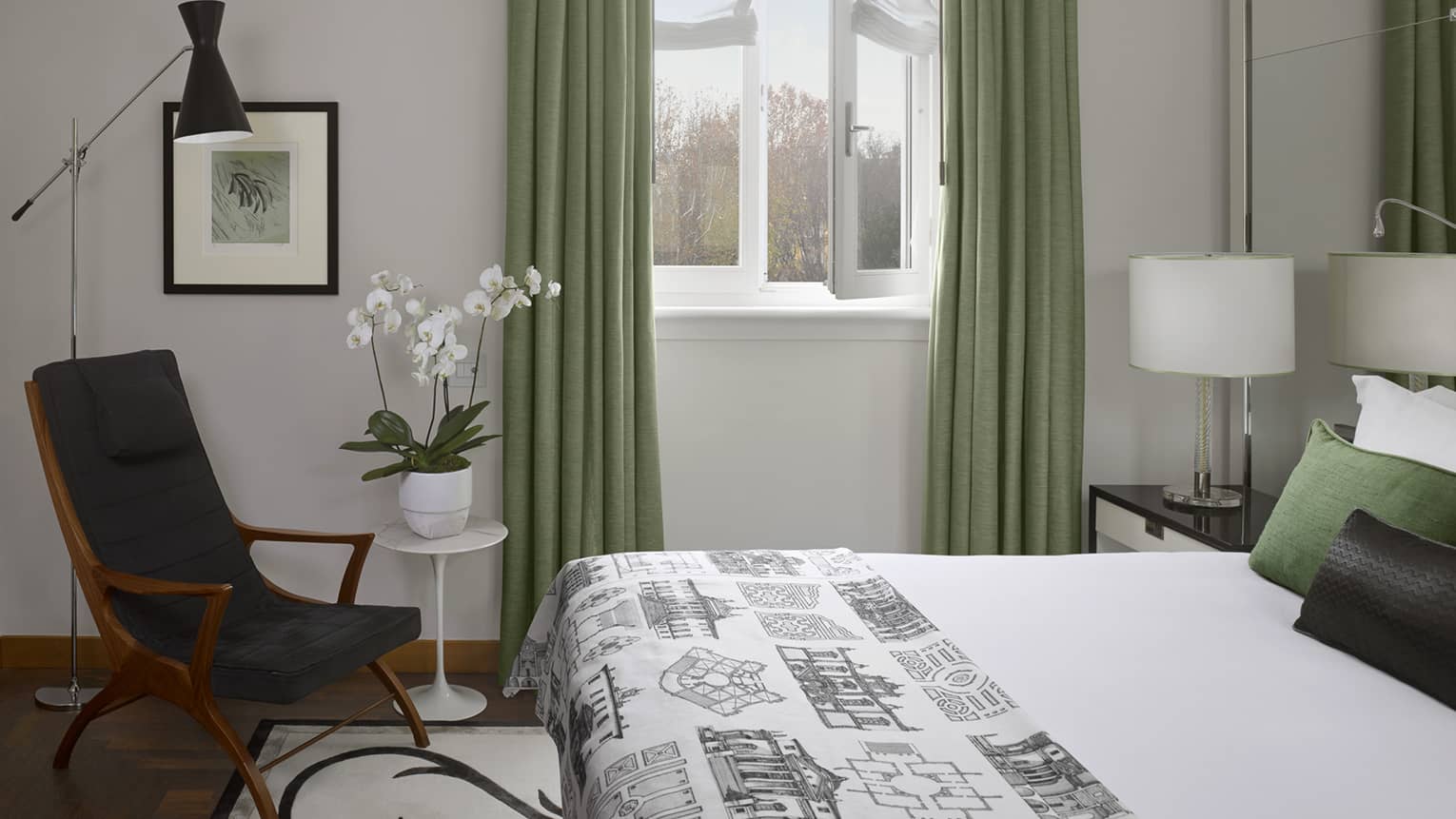 Hotel suite with white bed with graphic coverlet at foot and green-toned pillows beside window flanked with green curtains and modern armchair